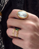 A hand wearing an oval ring with a grey shell stone,  a textured gold band ring and a ring with an emerald.