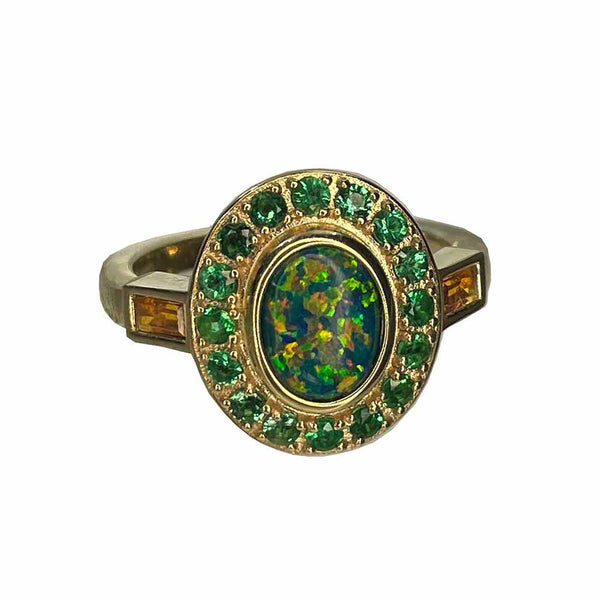 A gold ring with an oval opal center stone and green emerald halo of smaller stones, flanked by two citrine baguettes.
