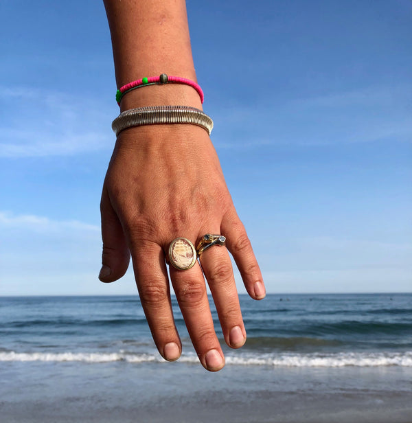 A hand in front of waves and blue sky wearing pink shell bracelet, silver bracelet and rings