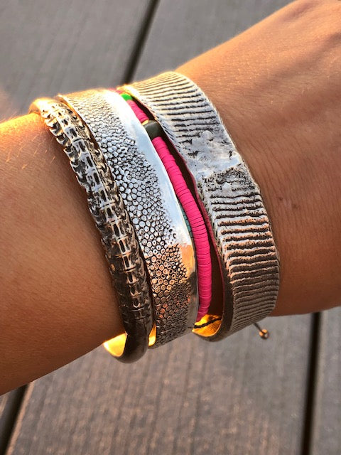 A group of silver nautical cuff bracelets on the wrist of a model.  There is a hot pink shell bracelet in the middle of the 3 silver bracelets.