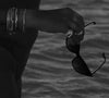 A black and white model shot of a hand holding sunglasses standing in the ocean.  The hand has silver rings on two fingers and silver bracelets worn on the wrist.