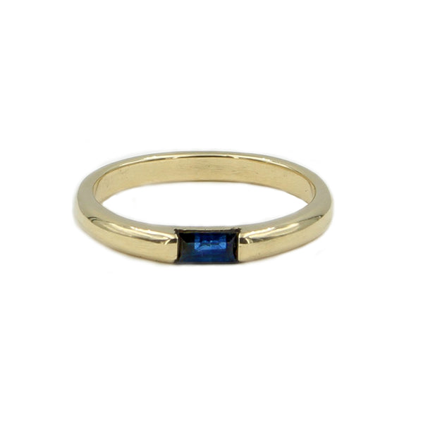 A gold band ring with a horizontal blue sapphire baguette set east to west in the middle.
