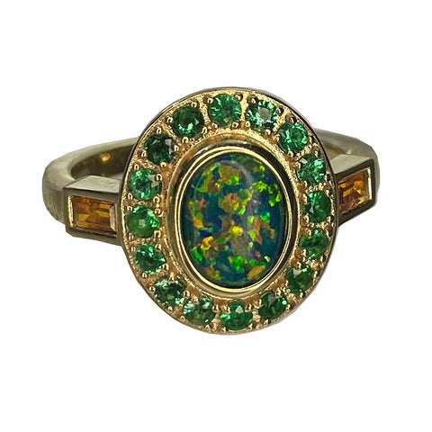 A gold ring with an oval opal center stone and green emerald halo of smaller stones, flanked by two citrine baguettes.