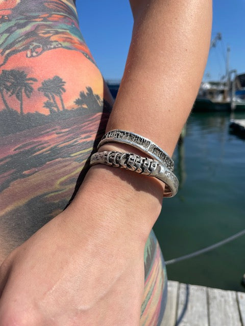 Two blackened silver cuff bracelets on a models arm.  The arm is resting on  tattoed skin and water and boats can be seen in the background.