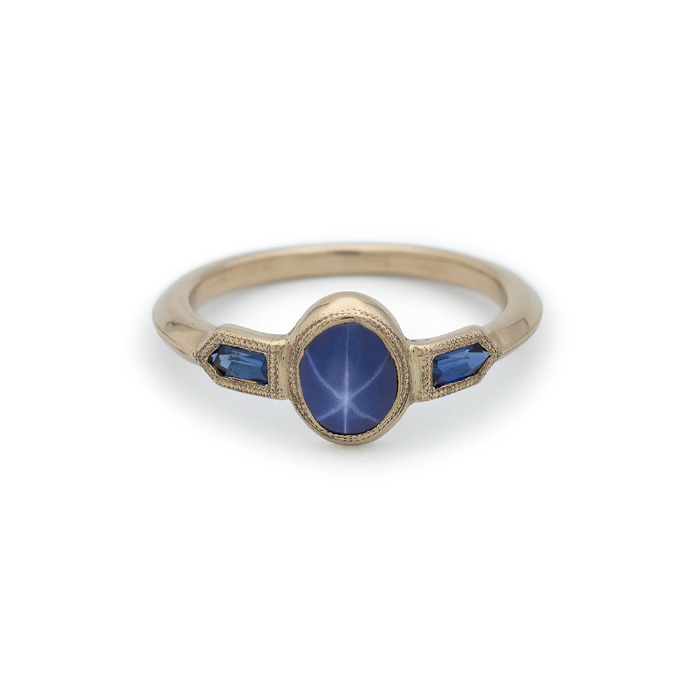 A gold ring with oval star sapphire in center and two bullet sapphires on sides with millgrain detail.